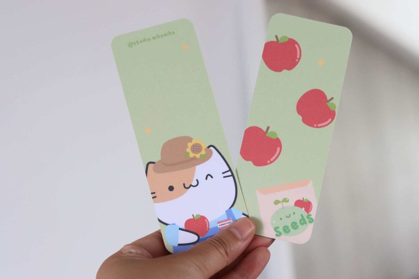 A hand holding a bookmark with an apple pattern and a bookmark with a cat in gardening clothes