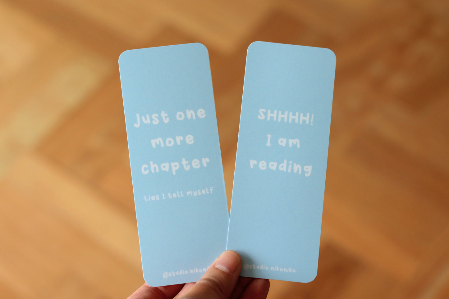 A hand holding two blue bookmarks with the texts: Just one more chapter and SHHH! I am reading