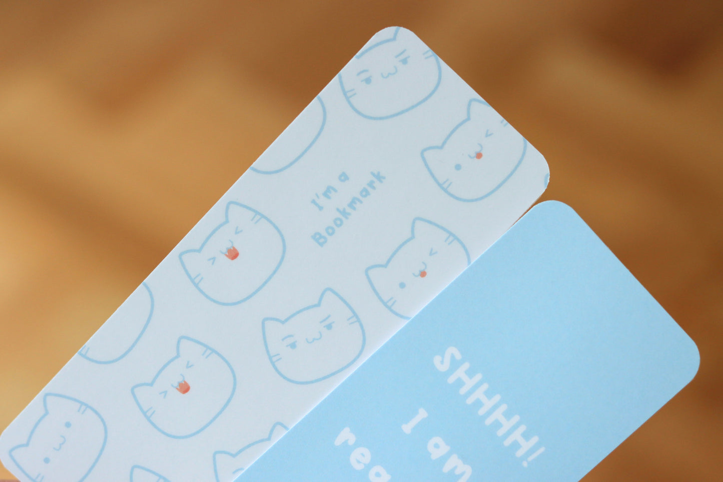 One white bookmark with a cat face pattern and the text (I am a bookmark) and a blue bookmark with text
