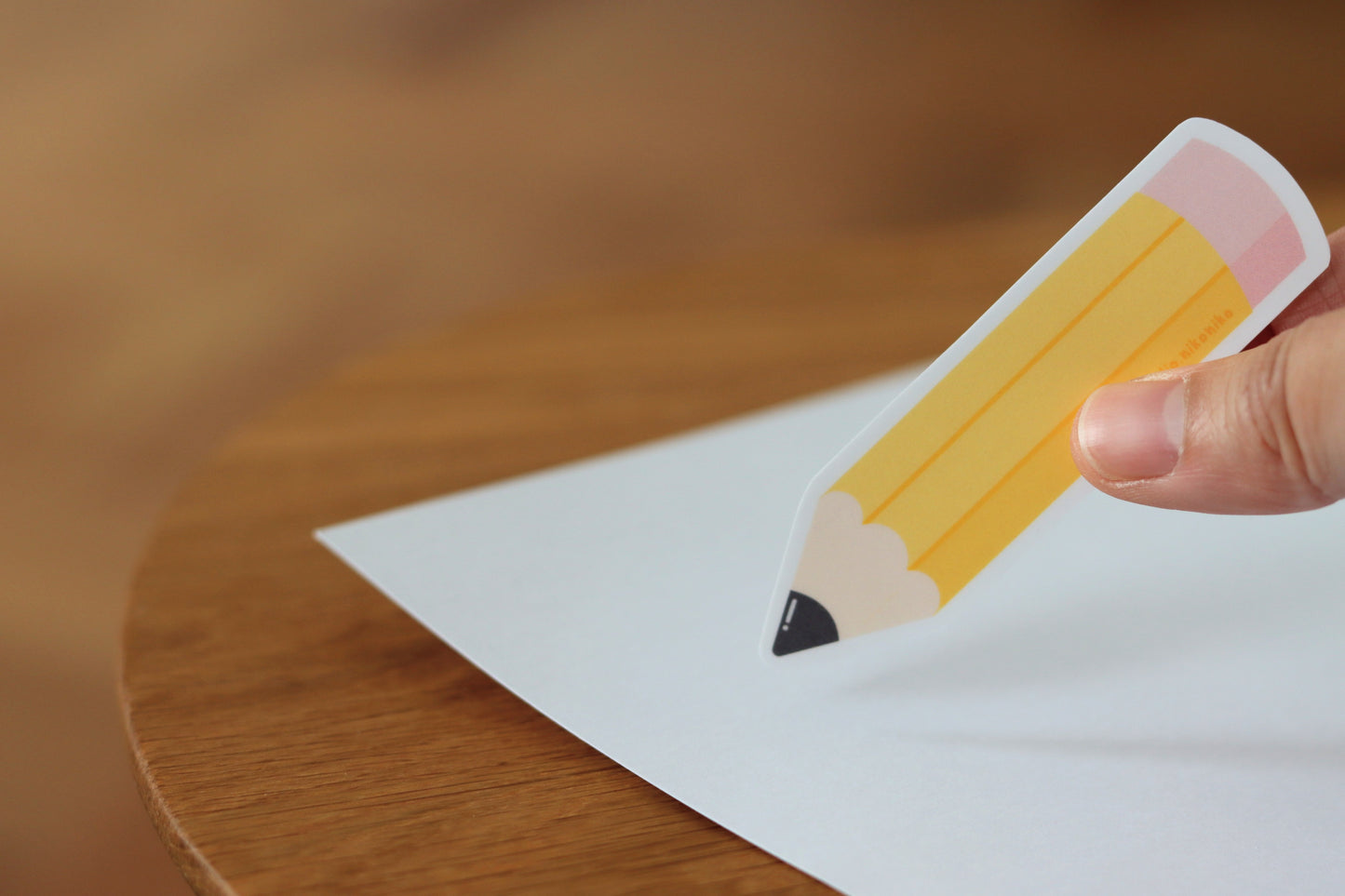 A hand holding a sticker design of a yellow pencil and pink eraser on a piece of white paper on a wooden table