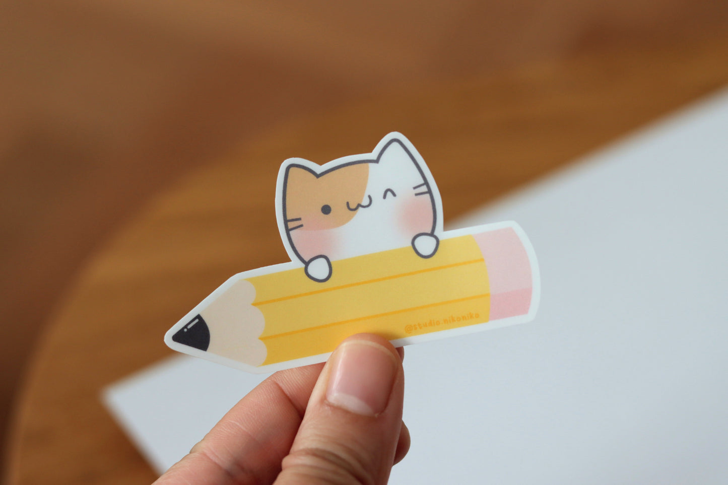 A hand holding a sticker design of a yellow pencil and pink eraser with a blushing cat