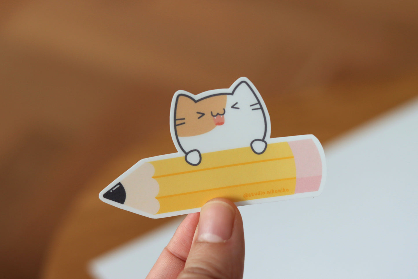 A hand holding a sticker design of a yellow pencil and pink eraser with a smiling cat